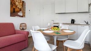 Un coin repas avec une table ronde, des chaises blanches, un canapé rouge et une cuisine en arrière-plan.