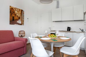 Un coin repas avec une table ronde, des chaises blanches, un canapé rouge et une cuisine en arrière-plan.