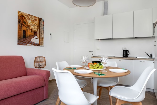 A dining area with a round table, white chairs, a red sofa, and a kitchen in the background.