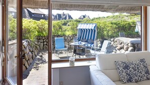 A cozy room with a large window, a white sofa, and a view of a garden with a striped awning and stone wall. - Herzlich Willkommen im Appartement Nepomuk (List auf Sylt)
