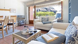 A living room with a sofa, a coffee table, and a TV. There is a window with a view of a house and trees. - Herzlich Willkommen im Appartement Nepomuk (List auf Sylt)