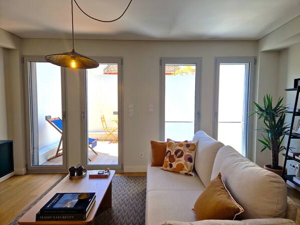 A living room with a sofa, a coffee table with books, a hanging light fixture, and a view through the glass doors.