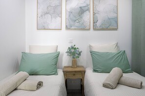 Quarto com duas camas, cada uma com um travesseiro verde e um travesseiro branco, uma mesa de cabeceira com um vaso de folhas verdes e três quadros abstratos na parede.
