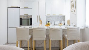 Una cocina moderna con armarios blancos, horno integrado y salpicadero de mármol. Hay cuatro sillas con cojines blancos y patas de madera.