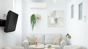 Una sala de estar moderna con un sofá beige, dos mesas redondas de centro, un televisor montado en la pared, un aire acondicionado y un estante con plantas.