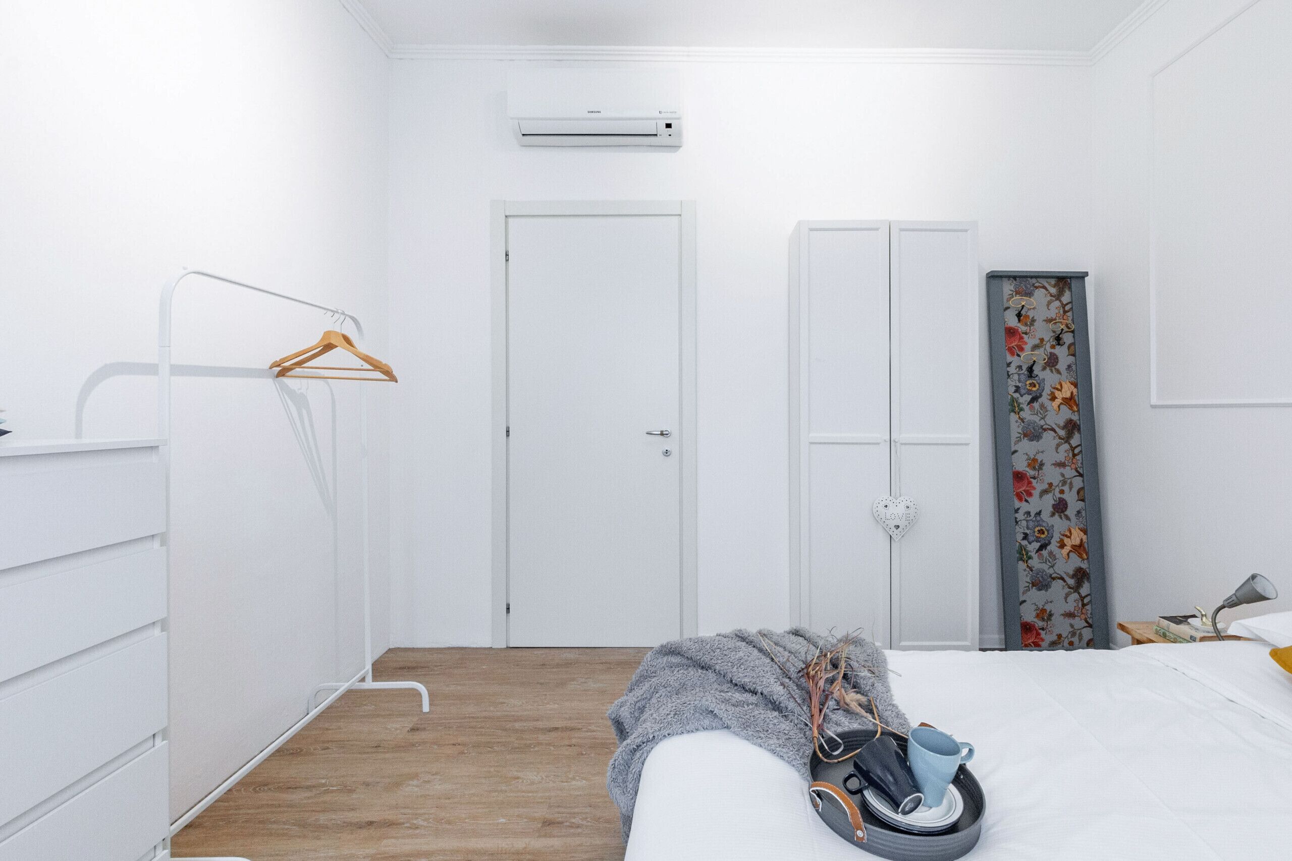 Un dormitorio minimalista con suelo de madera, una cama con manta gris, un zapatero colgado, un armario y una puerta con estampado floral.