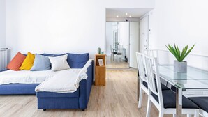 A modern living room with a blue sofa, white and colorful cushions, a glass dining table, white chairs, and a potted plant. - Golfo Aranci (Milano)