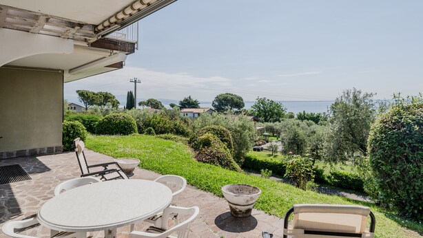 En terrasse med et bord og stole, en potteplante og udsigt til havet og grønne omgivelser.