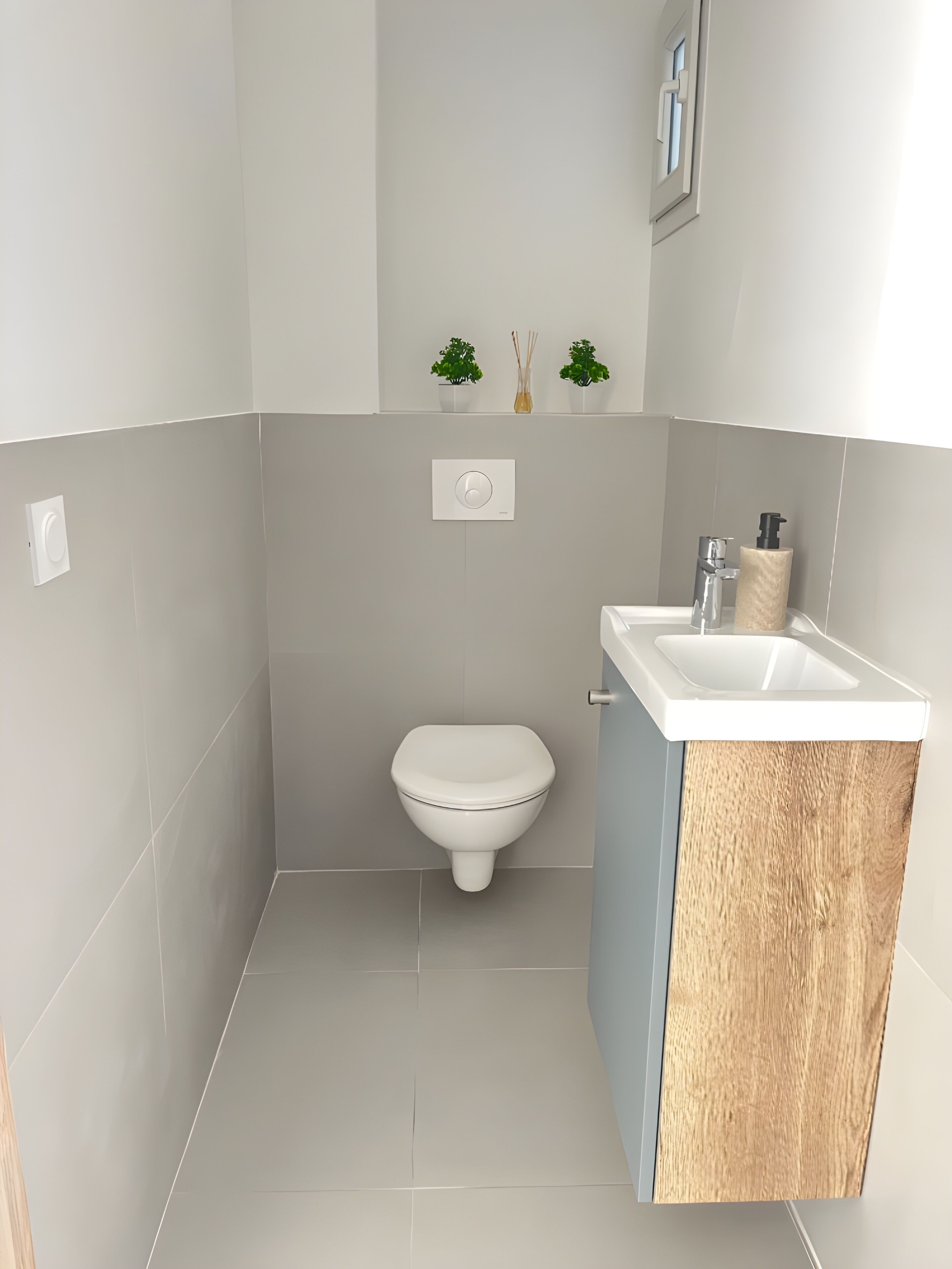A modern bathroom with a toilet, a sink with a wooden cabinet, and two small potted plants on the wall.