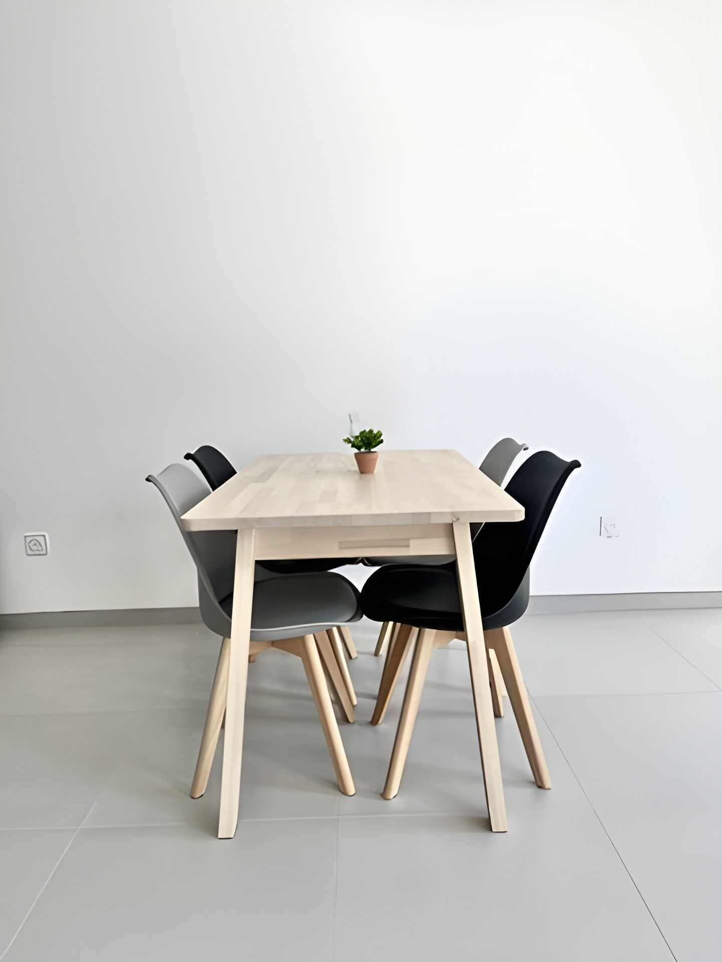 Taula rectangular de fusta amb quatre cadires negres, una petita planta en test i una paret blanca.