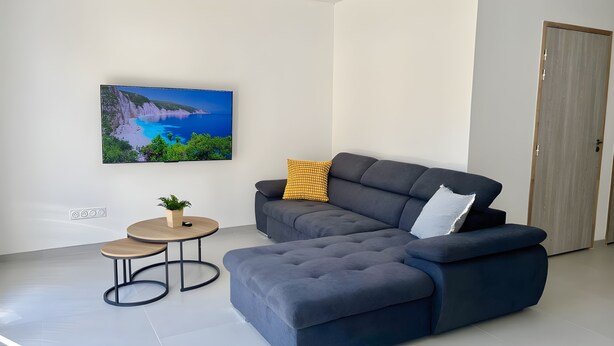 Een moderne woonkamer met een grijze modulaire bank, een klein rond salontafeltje en een flatscreen televisie aan de muur.