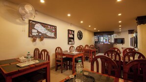 A dining area with wooden tables and chairs, a wall-mounted fan, and a bar area in the background. - Hotel Nomad Inn (Pokhara)