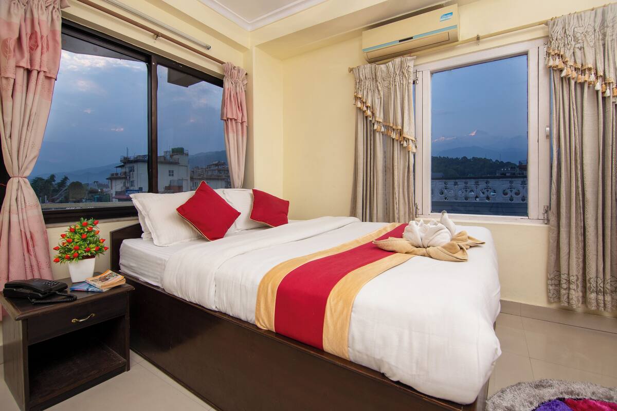 A hotel room with a large bed, a bedside table with a phone and a plant, and a window with curtains.