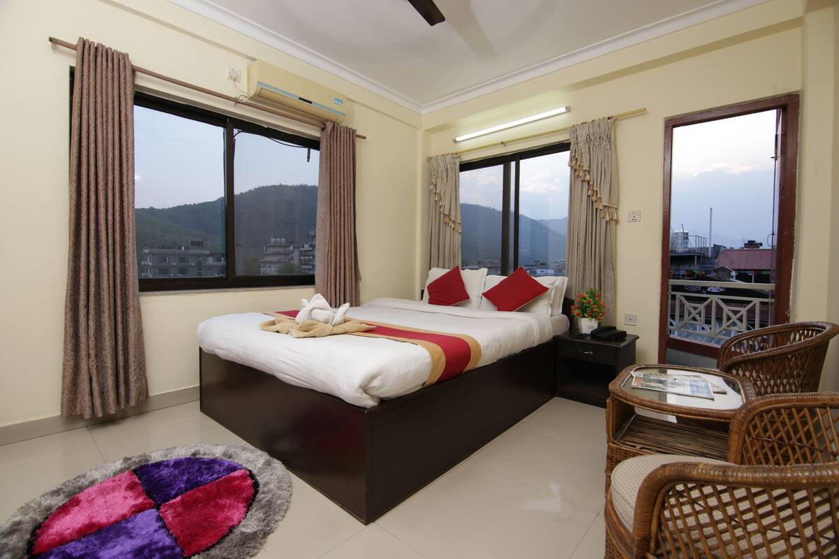 A hotel room with a large bed, two windows with curtains, a small table with chairs, and a view of the city and mountains.