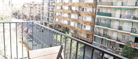Un balcón con banca de madera, con vista a una calle flanqueada por edificios de varios pisos.