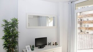 A modern room with a large window, a flat-screen TV on a white cabinet, a mirror above the TV, and a potted plant to the left. - Sant Jordi Apartments Sagrada Familia (Barcelona)