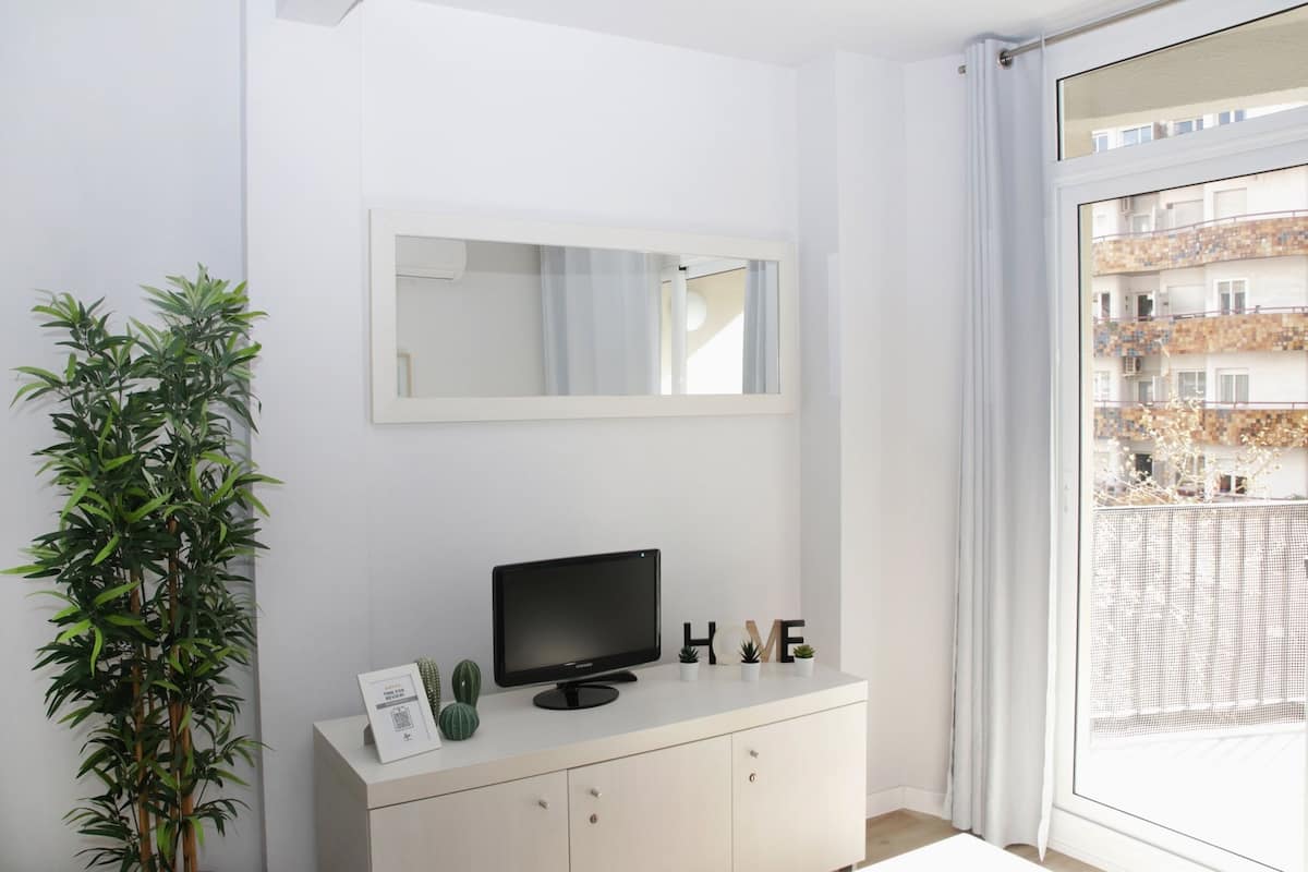 A modern room with a large window, a flat-screen TV on a white cabinet, a mirror above the TV, and a potted plant to the left.