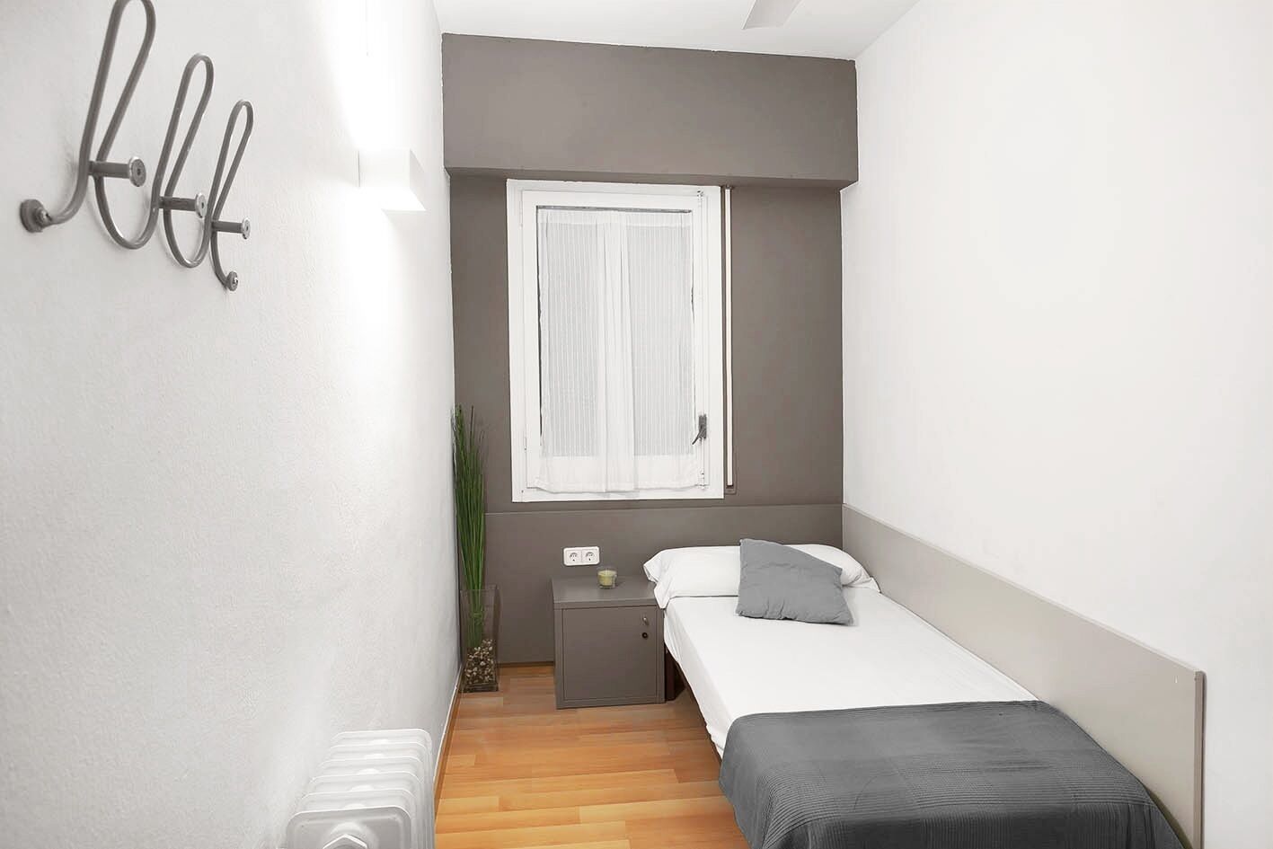 Un dormitorio pequeño y moderno con una cama individual, una mesita de noche, cortinas blancas en una ventana y un aplique de pared.