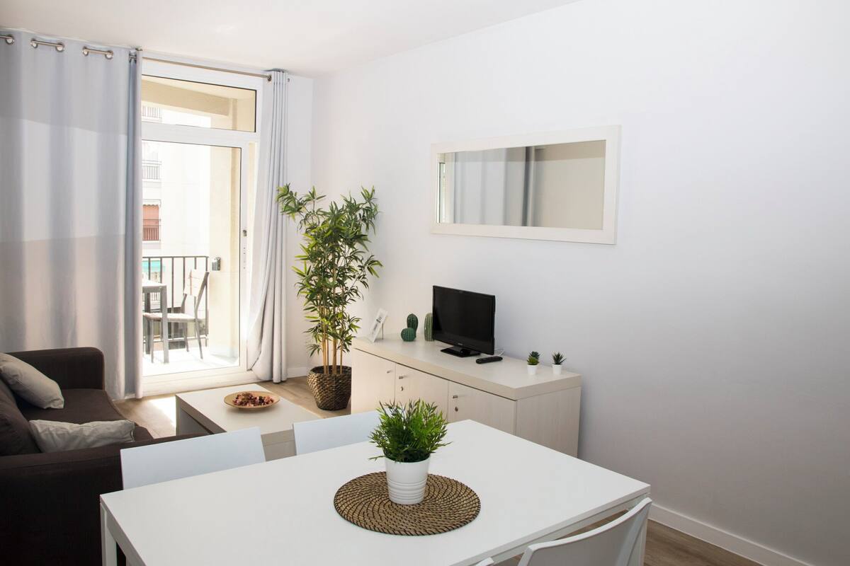 A modern living room with a sofa, a dining table with a plant, a TV on a cabinet, and a balcony with a view.
