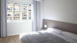 A hotel room with a large bed, a nightstand, a window with curtains, and a view of other buildings. - Sant Jordi Apartments Sagrada Familia (Barcelona)