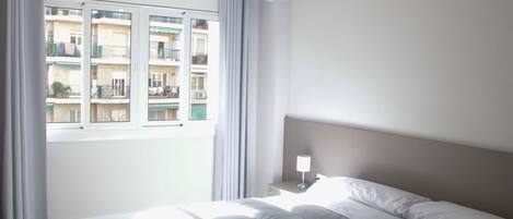 Habitación de hotel con una cama grande, una mesita de noche, una ventana con cortinas y vistas a otros edificios.