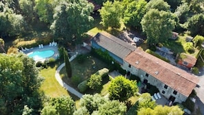 An aerial view of a property with a swimming pool, a large building with a tiled roof, and surrounding greenery.