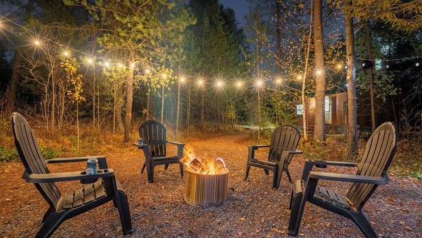 A cozy outdoor seating area with Adirondack chairs around a campfire, surrounded by trees and string lights.