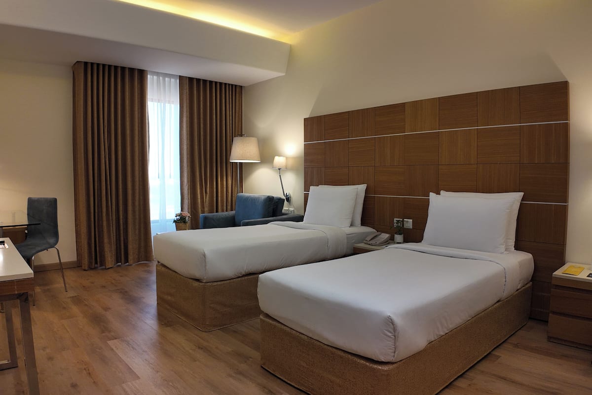 A hotel room with two beds, a desk, a chair, a lamp, and a window with curtains.
