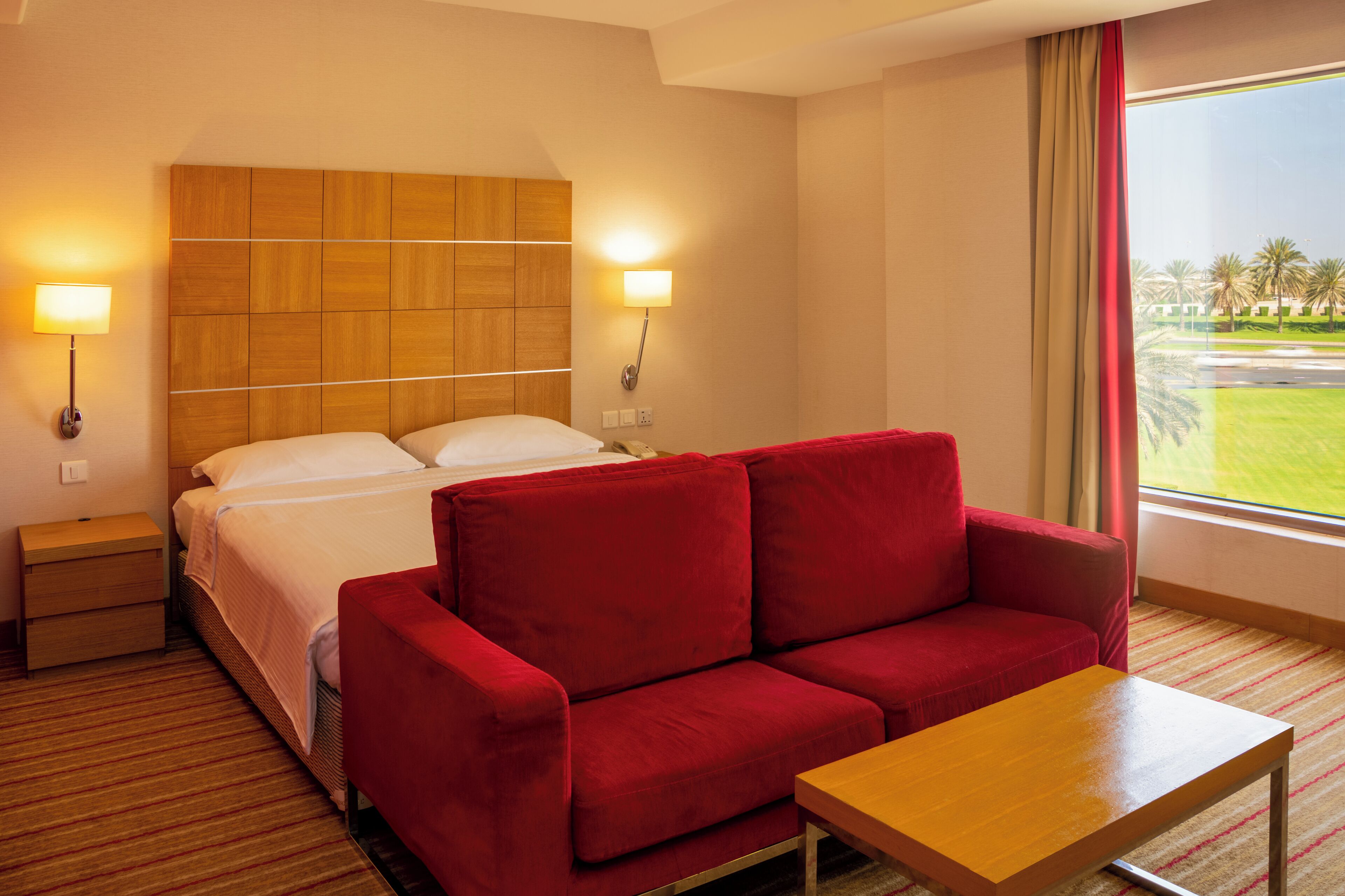 Una camera d'albergo con un letto, un divano rosso, un comodino in legno, un tavolo in legno e un'ampia finestra con vista su un campo da golf.