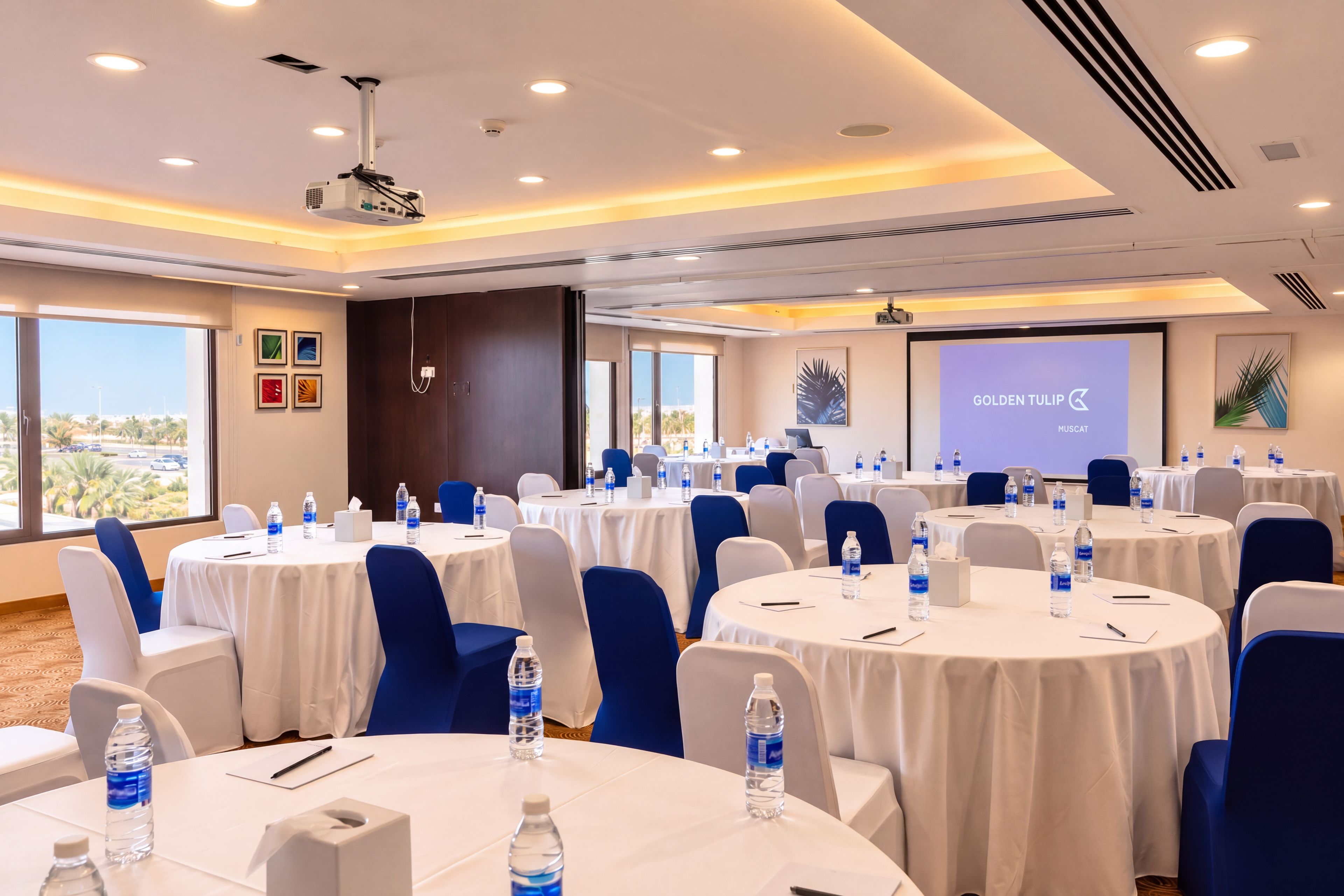 A conference room with multiple tables, chairs, and a projector screen displaying 'GOLDEN TULIP MUSCAT'.