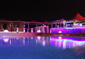 Zona de piscina nocturna con bar y asientos bajo toldos rojos.