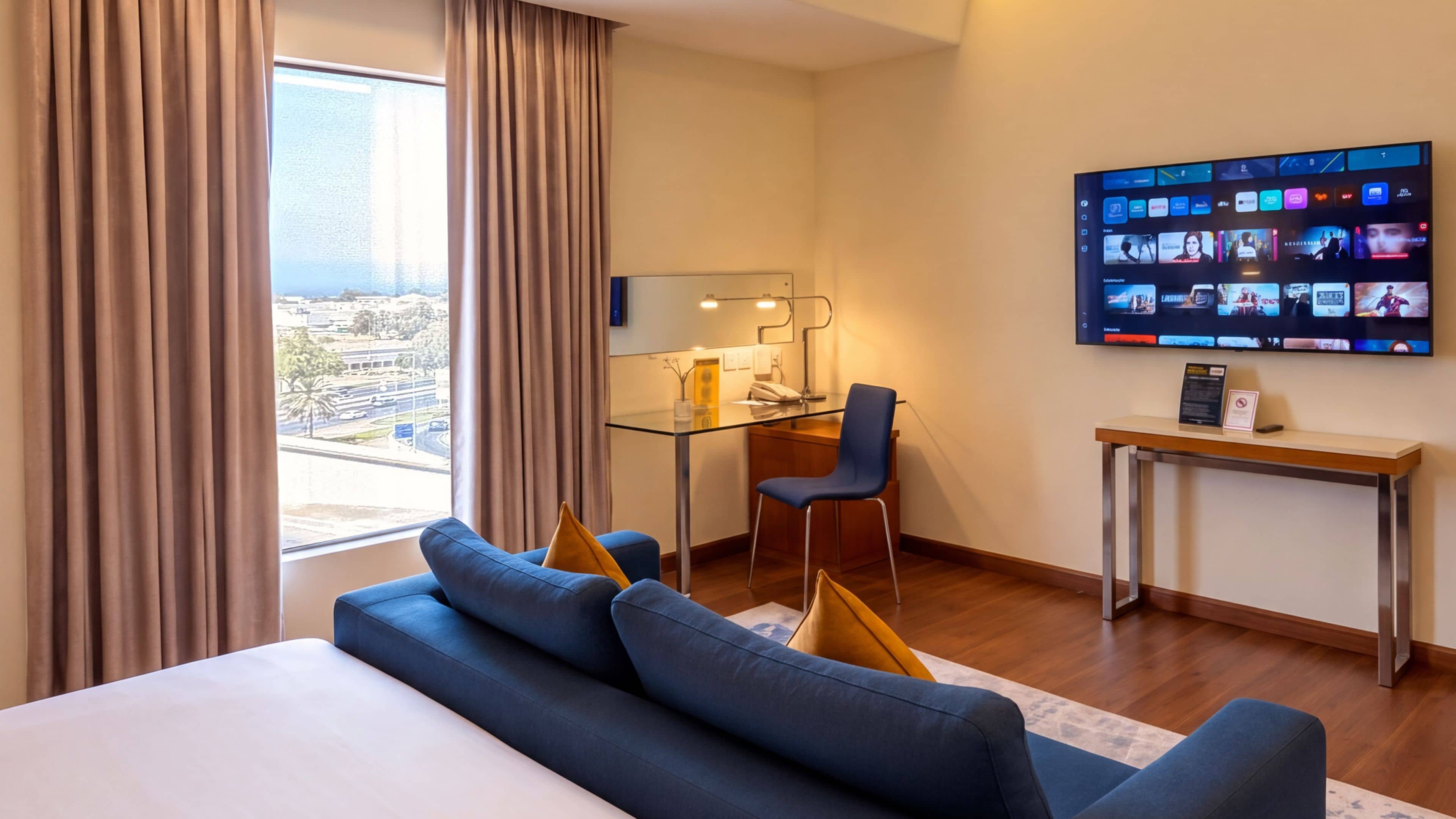 A hotel room with a large bed, a desk with a chair, a TV displaying various channels, and a view of the city through the window.