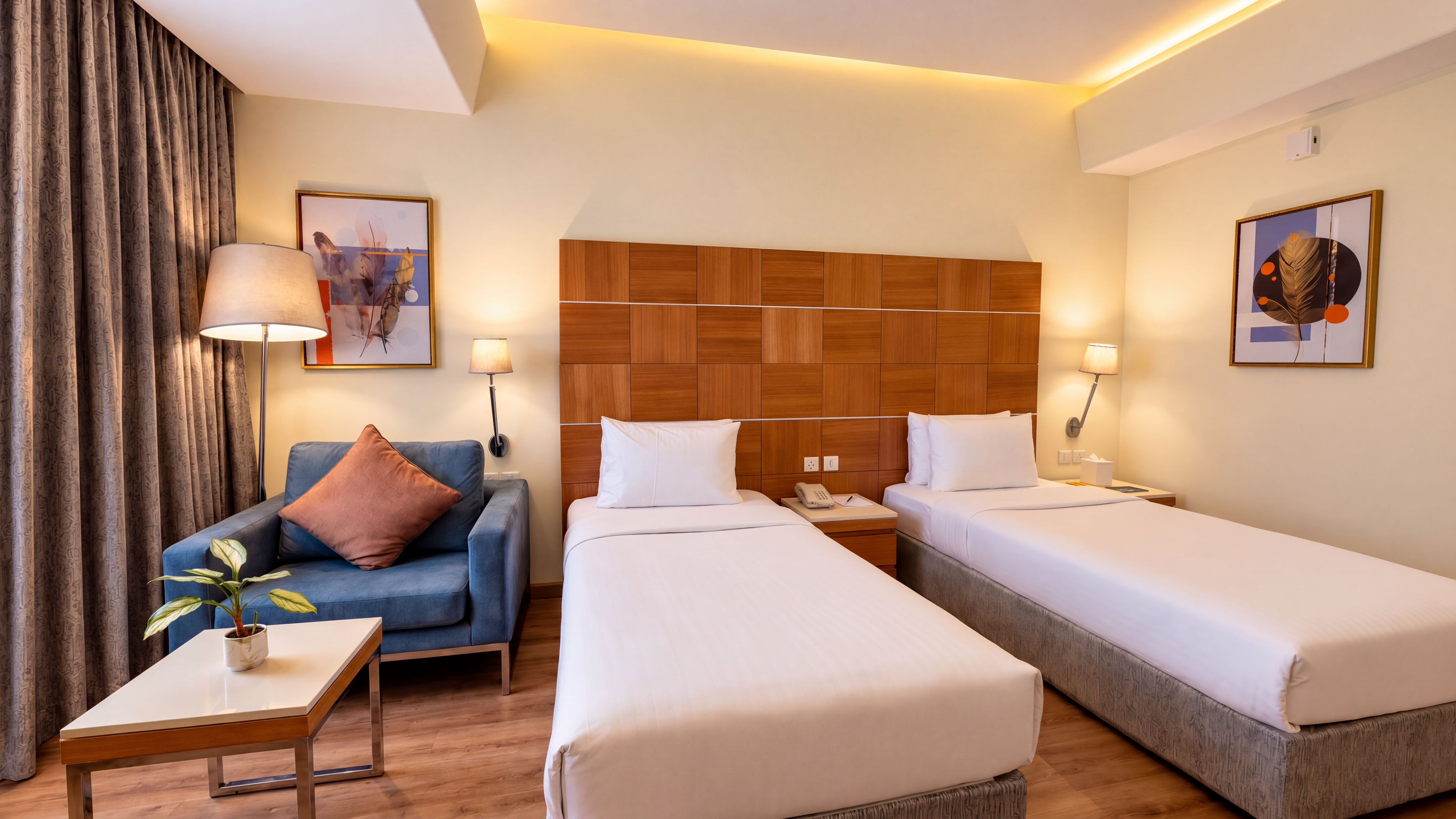 A hotel room with a wooden headboard, two single beds, a blue sofa, a small table with a plant, and wall-mounted lamps.