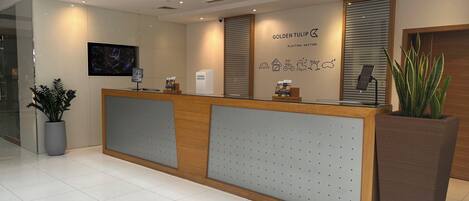 A hotel reception desk with a modern design, a large potted plant, and a wall-mounted screen displaying a cityscape.