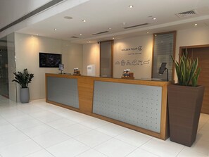 A hotel reception desk with a modern design, a large potted plant, and a wall-mounted screen displaying a cityscape.