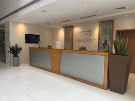 A hotel reception desk with a modern design, a large potted plant, and a wall-mounted screen displaying a cityscape.