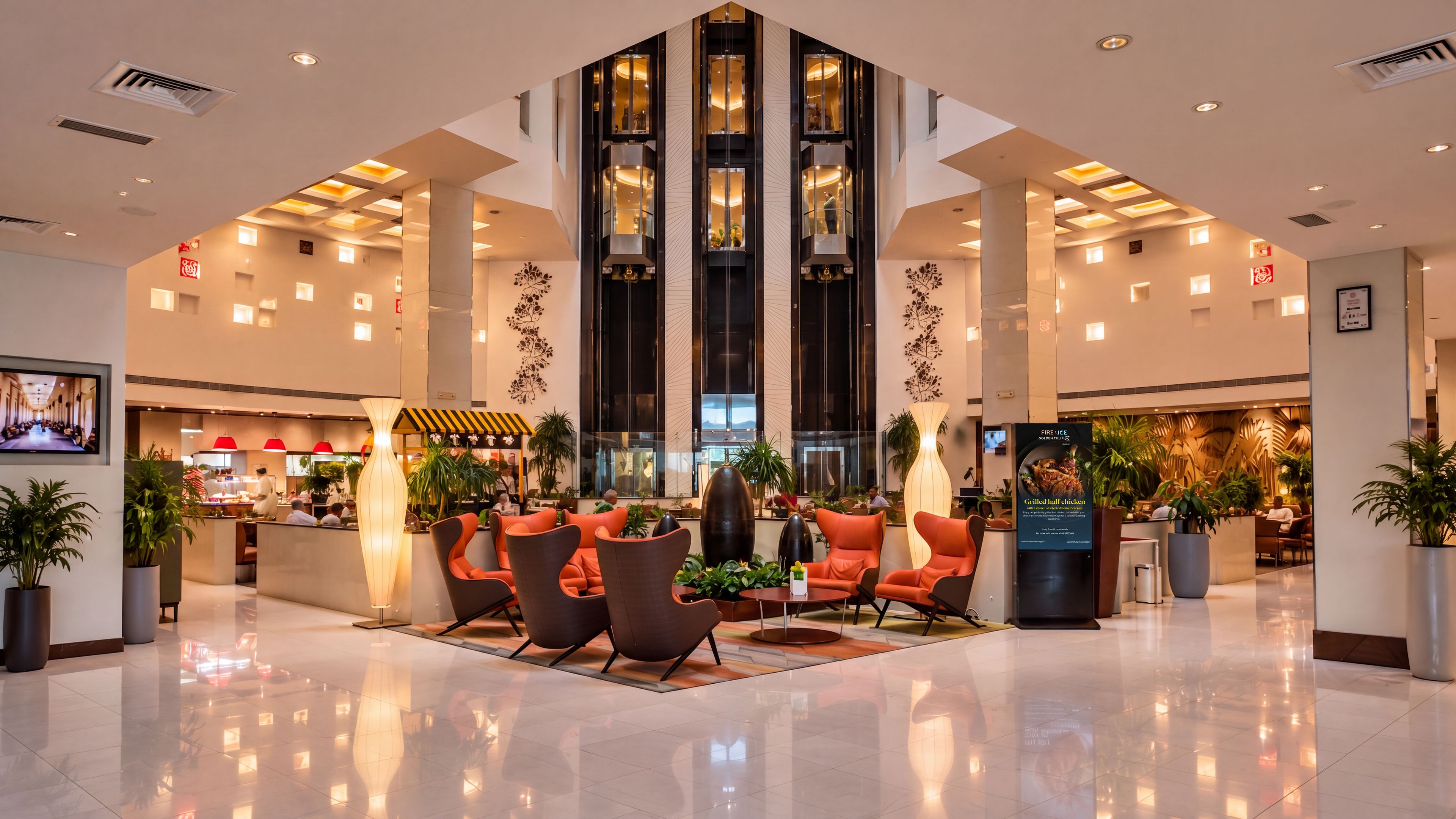 A modern hotel lobby with a central seating area, a restaurant in the background, and a large digital screen.