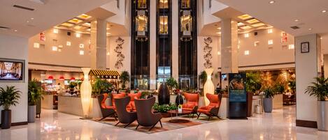 A modern hotel lobby with a central seating area, a restaurant in the background, and a large digital screen.
