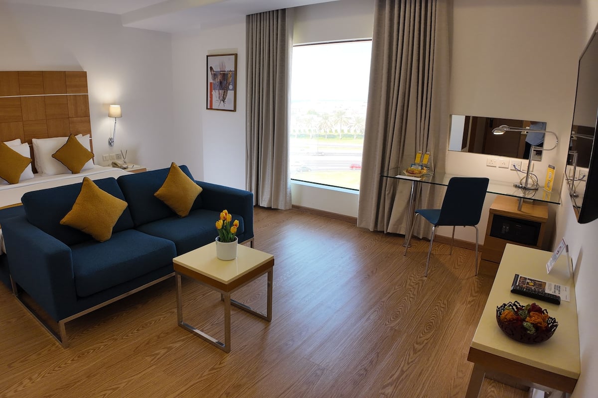 A hotel room with a bed, a blue sofa, a small table with a vase of flowers, a desk with a chair, and a large window with curtains.