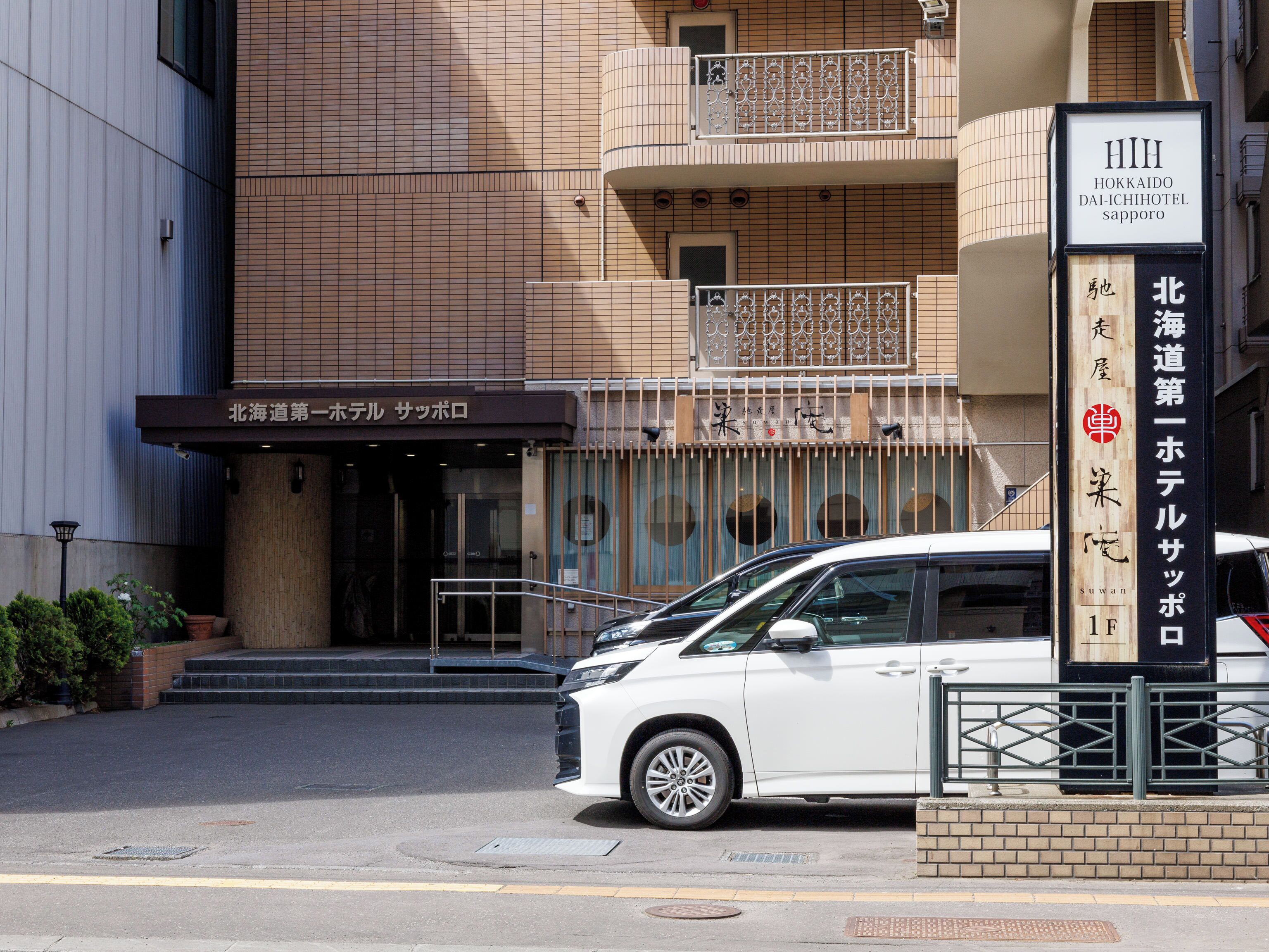 Entrada de un hotel con un cartel en japonés, un coche blanco estacionado al frente y un edificio de fachada con tejas.