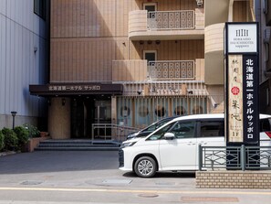 Entrada de un hotel con un cartel en japonés, un coche blanco estacionado al frente y un edificio de fachada con tejas.
