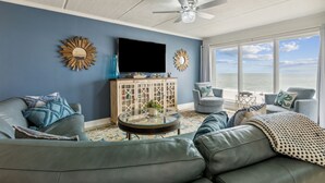 A living room with a large TV, two sofas, a glass coffee table, and a view of the ocean.