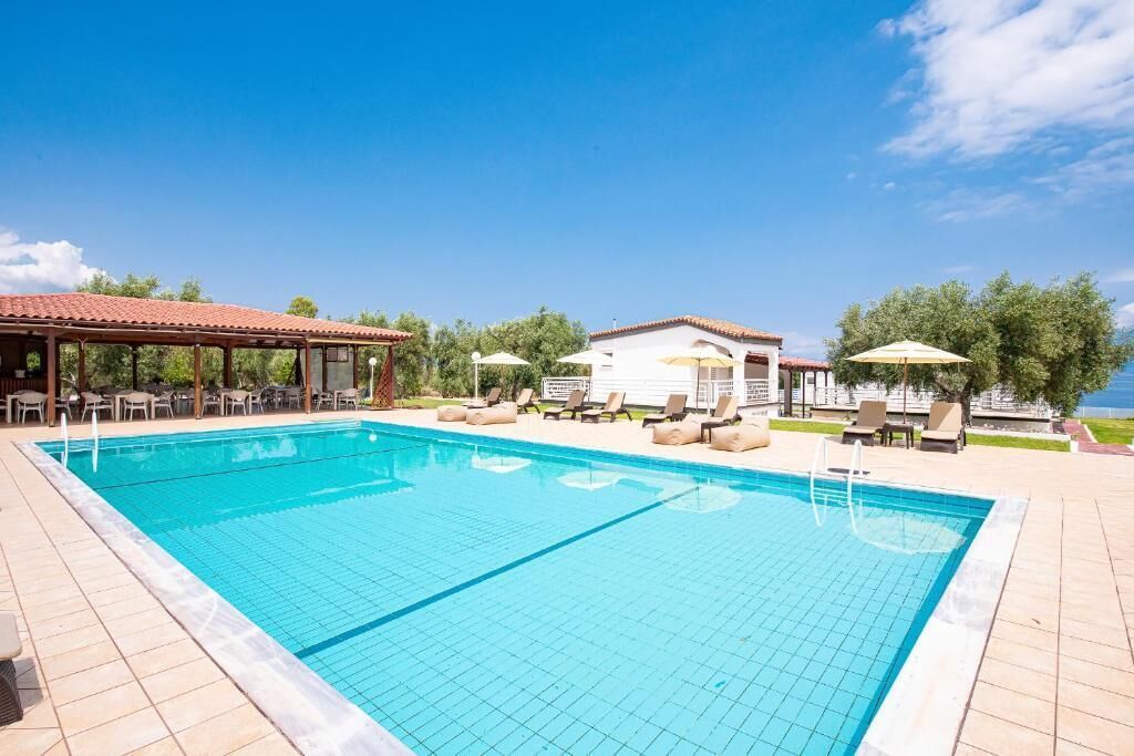 A swimming pool with lounge chairs and umbrellas, surrounded by a covered seating area and a building with a tiled roof.