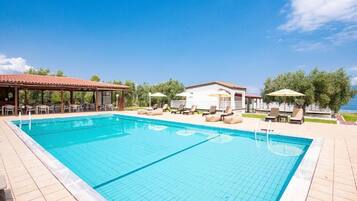 A swimming pool with lounge chairs and umbrellas, surrounded by a covered seating area and a building with a tiled roof.