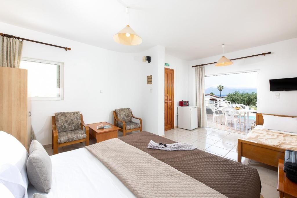 A hotel room with a bed, two armchairs, a small table, a TV, and a balcony with outdoor seating.