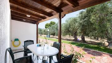 A covered patio with a round table and chairs, overlooking a garden with trees and a pathway.