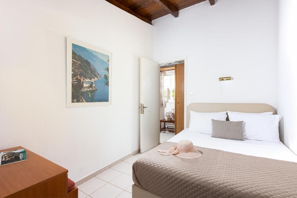 A bedroom with a bed, a wooden nightstand, a framed picture of a coastal landscape, and a door leading to another room.