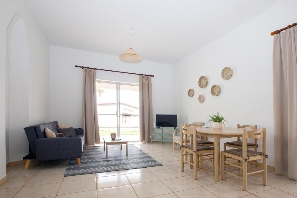 Una sala de estar con sofá, mesa de centro, mesa de comedor, sillas, televisor y un gran ventanal con cortinas.
