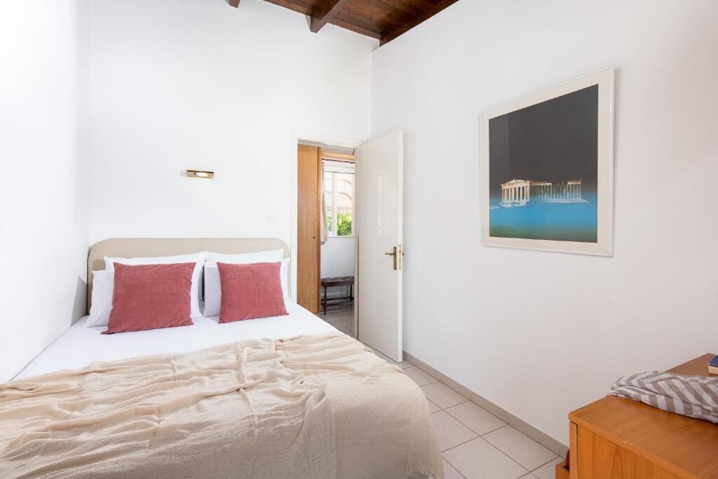 Een slaapkamer met een bed, een houten nachtkastje, een deur naar een andere kamer en een ingelijst kunstwerk aan de muur.