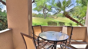 A patio with a round glass table and metal chairs, overlooking a grassy area with trees and a palm. - Catalina Foothills Condo on the Golf Course (Tucson)